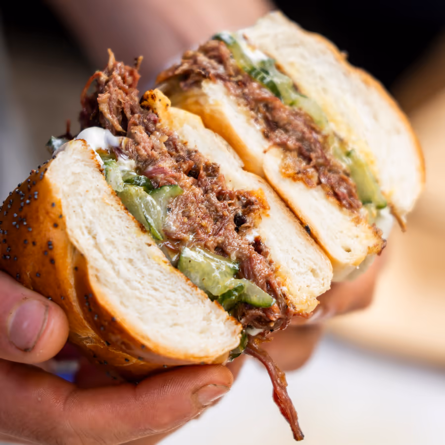 Hände halten ein aufgeschnittenes Brötchen mit Fleisch und Gurke