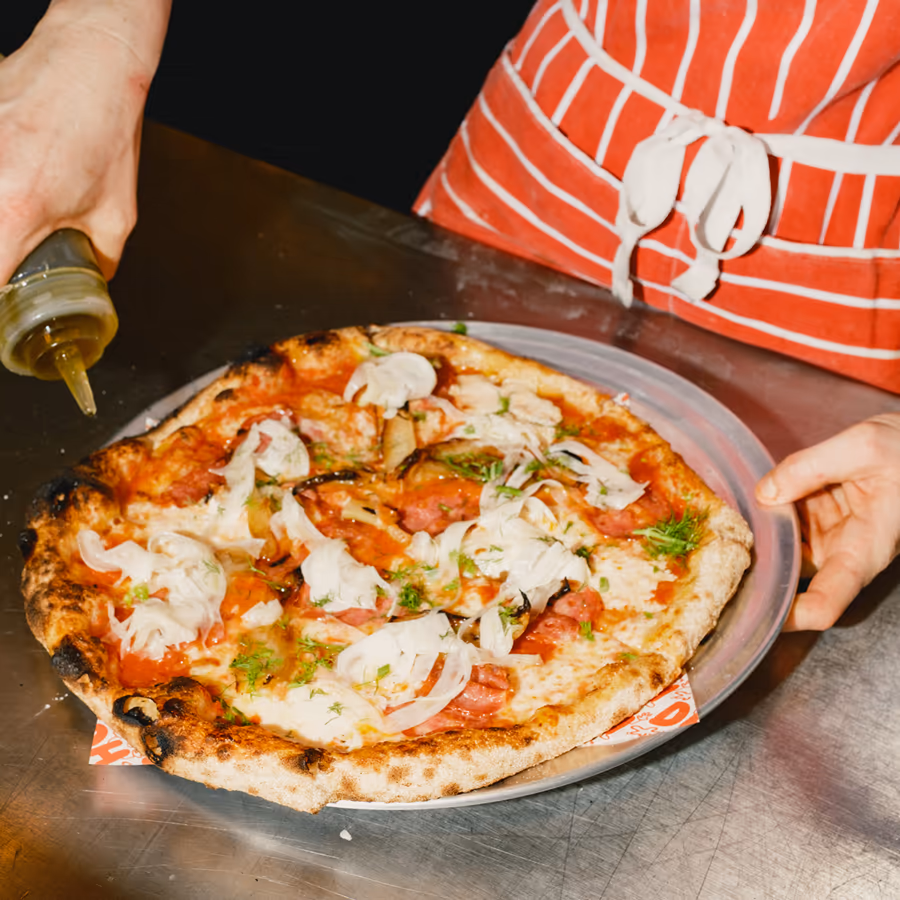Person beträufelt eine frisch belegte Pizza auf einem Metalltablett mit Öl